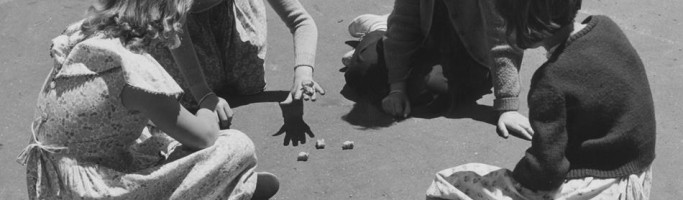 Photo from Museum Victoria and shows four young girls sitting in a circle on an asphalt surface, playing with sheep knucklebones in a government school playground in 1954.