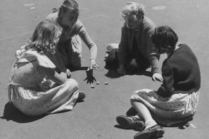 Photo from Museum Victoria and shows four young girls sitting in a circle on an asphalt surface, playing with sheep knucklebones in a government school playground in 1954.